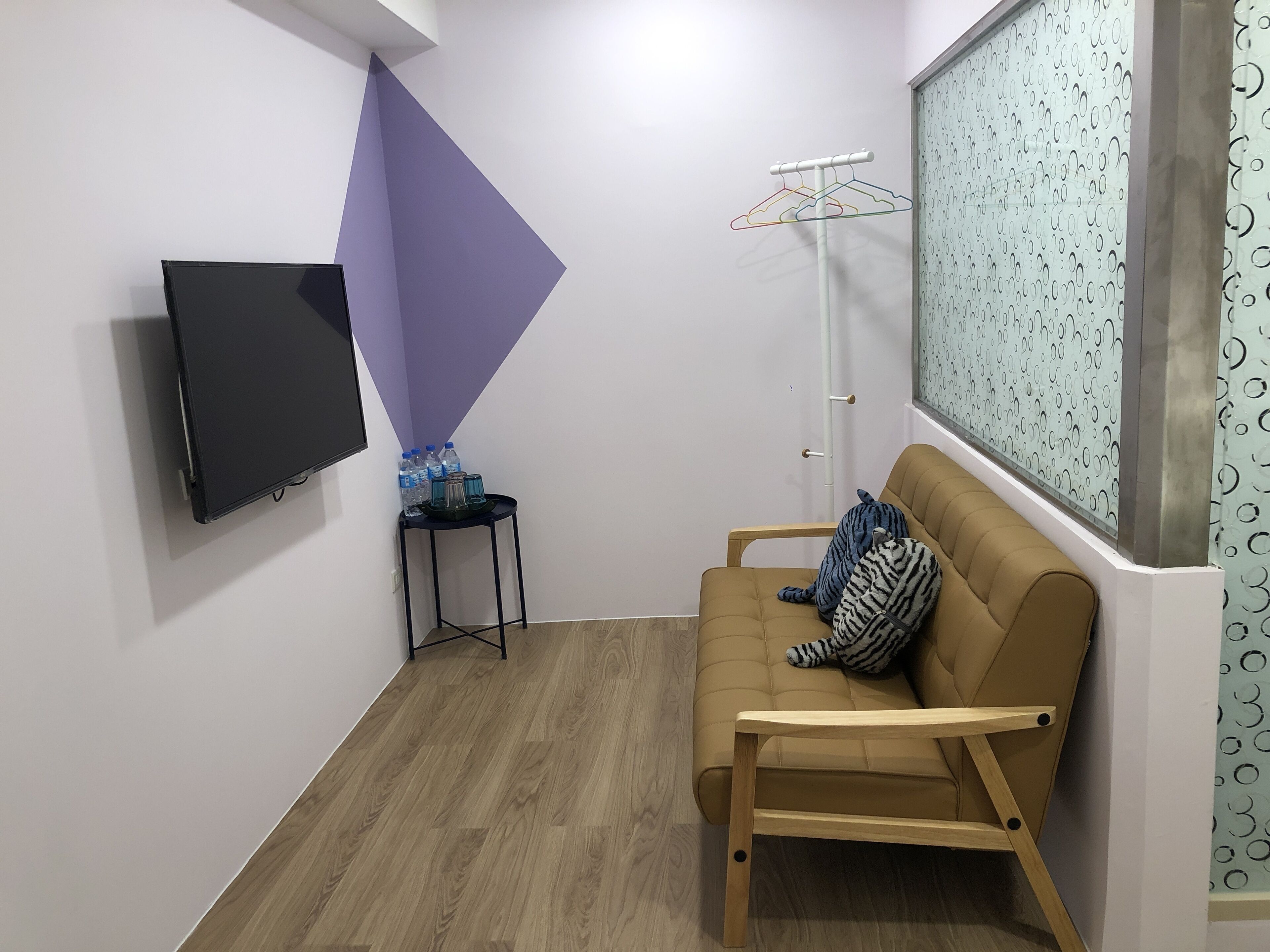 A room with a flat-screen TV mounted on the wall, a corner with a purple triangular accent, a small table with water bottles, a sofa with a striped pillow, and a window with a patterned curtain.