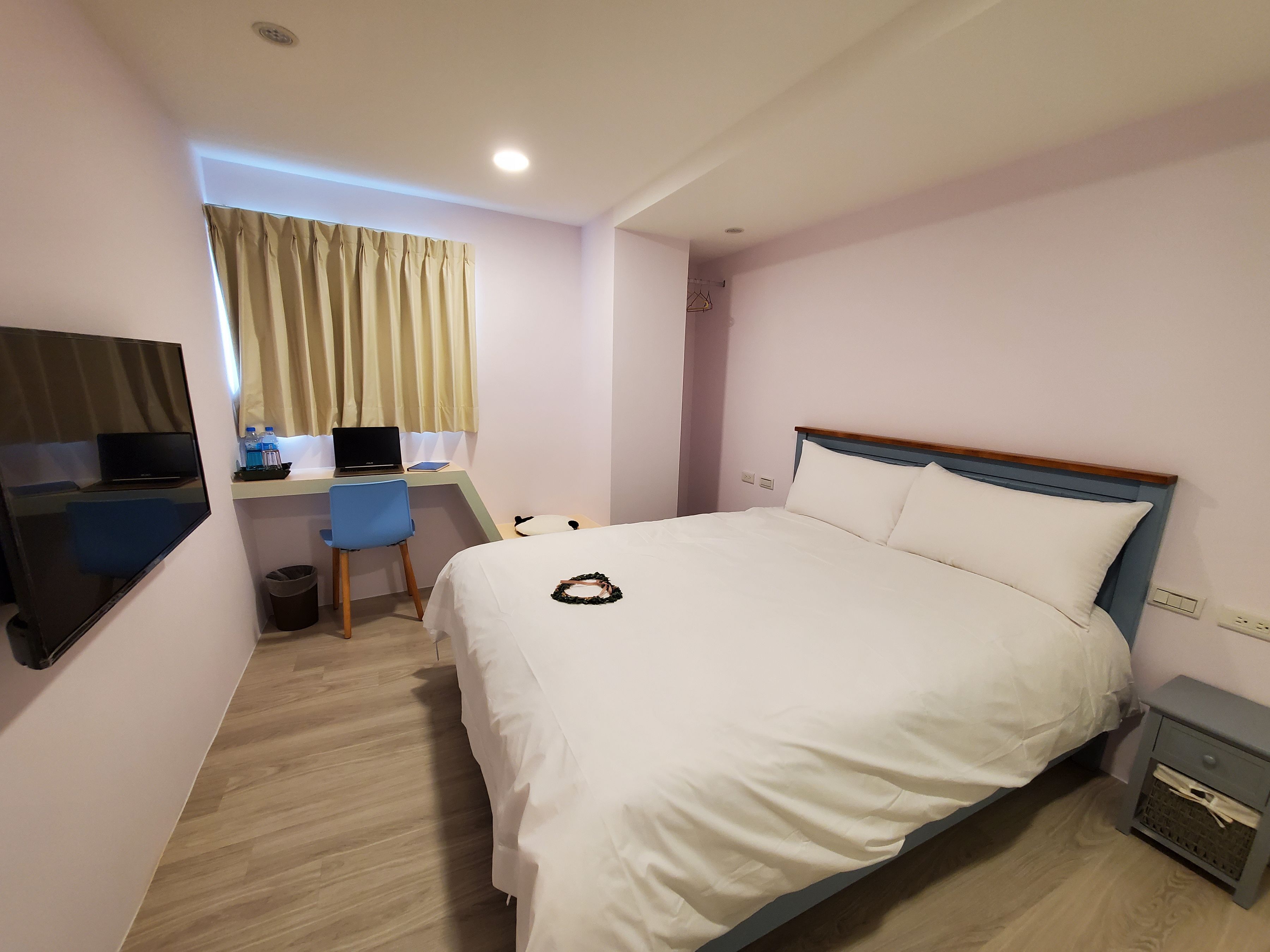 A hotel room with a bed, a desk with a laptop, a chair, a TV, and a small bedside table.