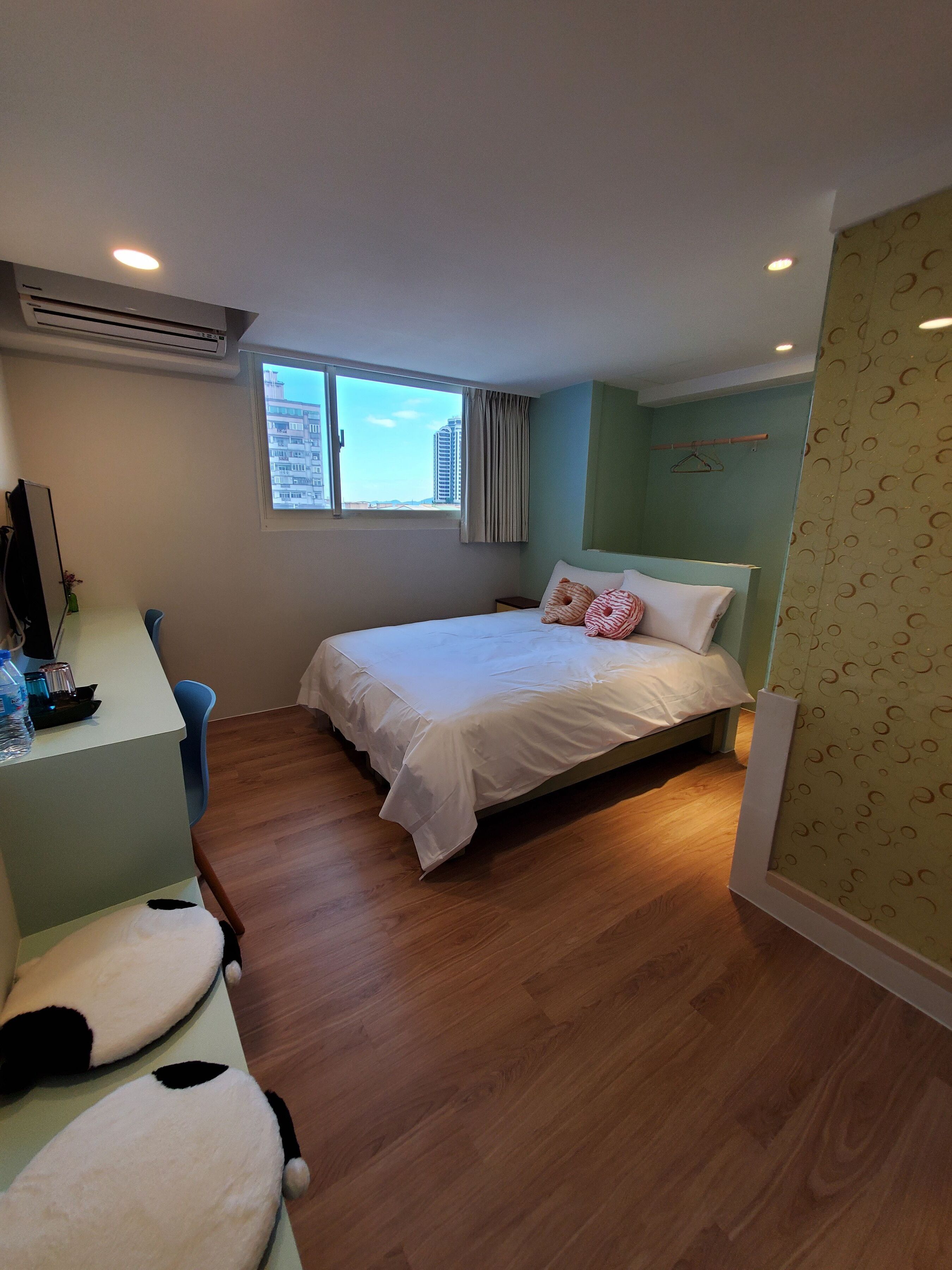 A bedroom with a bed, a desk, a chair, a hanging coat rack, and a window with a view of buildings.
