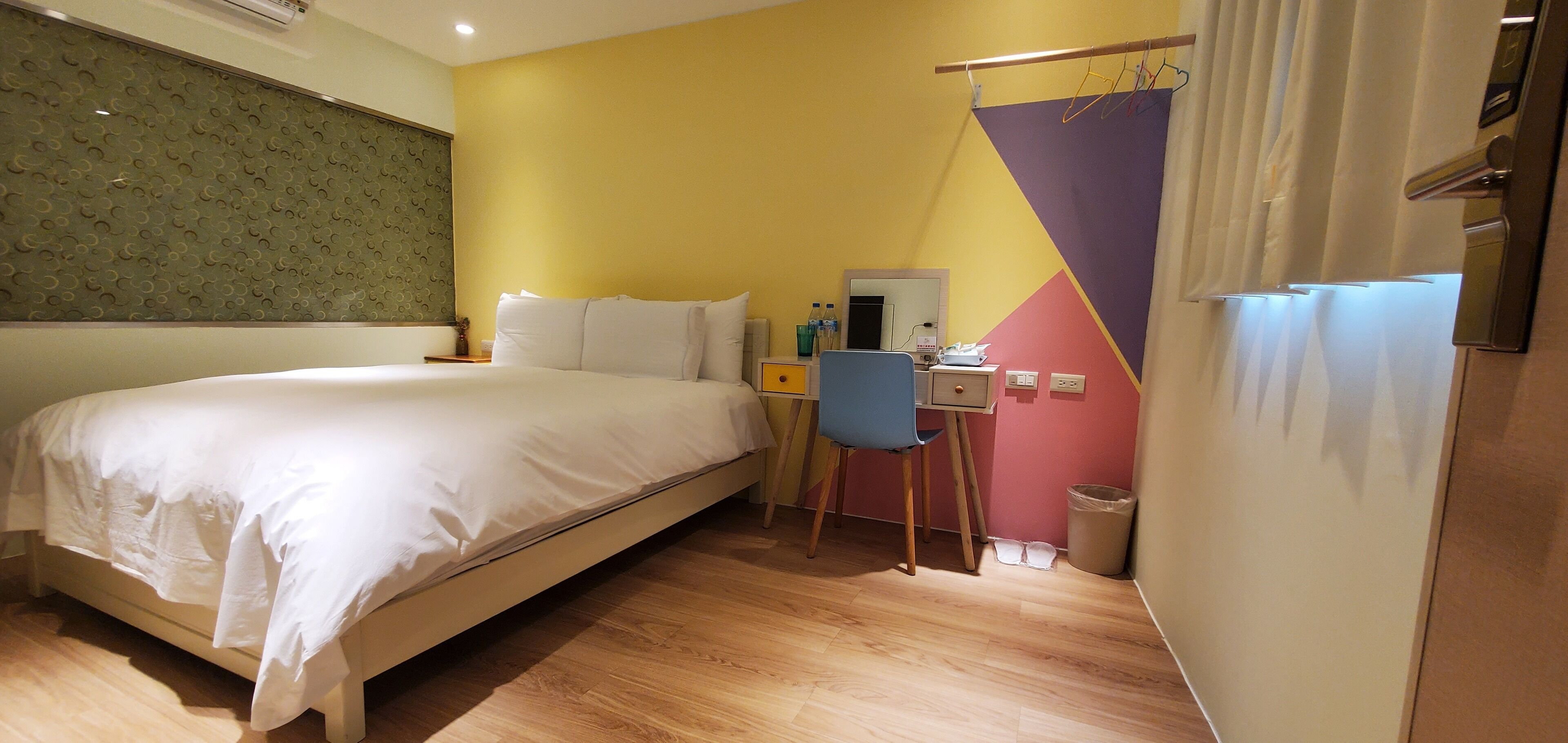 A hotel room with a bed, a desk, a chair, a mirror, and a trash bin.
