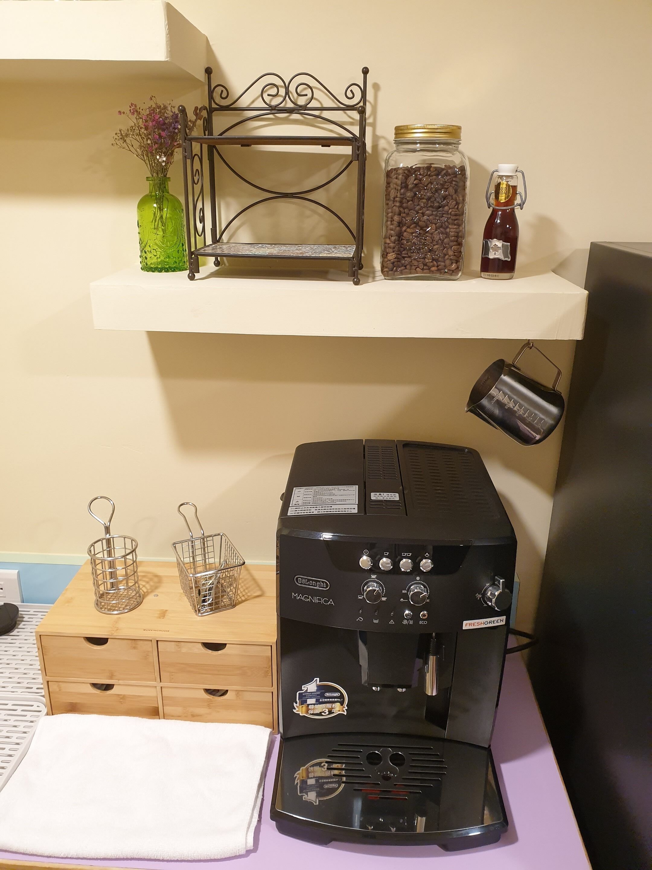 A coffee machine on a table with a shelf above it holding a metal rack, jars, and a bottle.