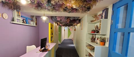Uma área de recepção com piso de madeira, paredes roxas e um teto decorado com arranjos florais pendentes. Há prateleiras com plantas e uma porta azul à direita.