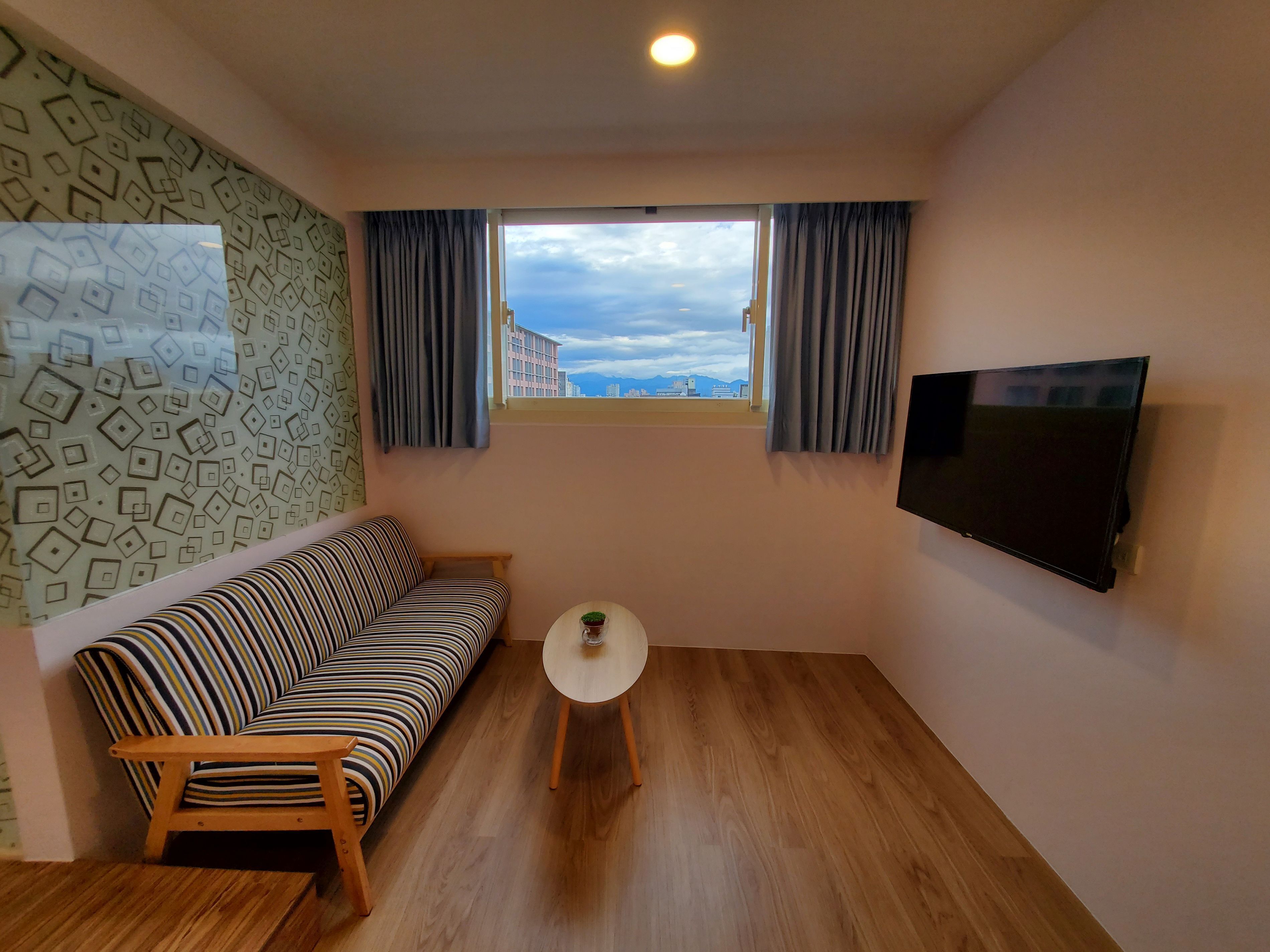 Un salón con un sofá a rayas, una mesita redonda con una planta, un televisor montado en la pared y una ventana con cortinas.