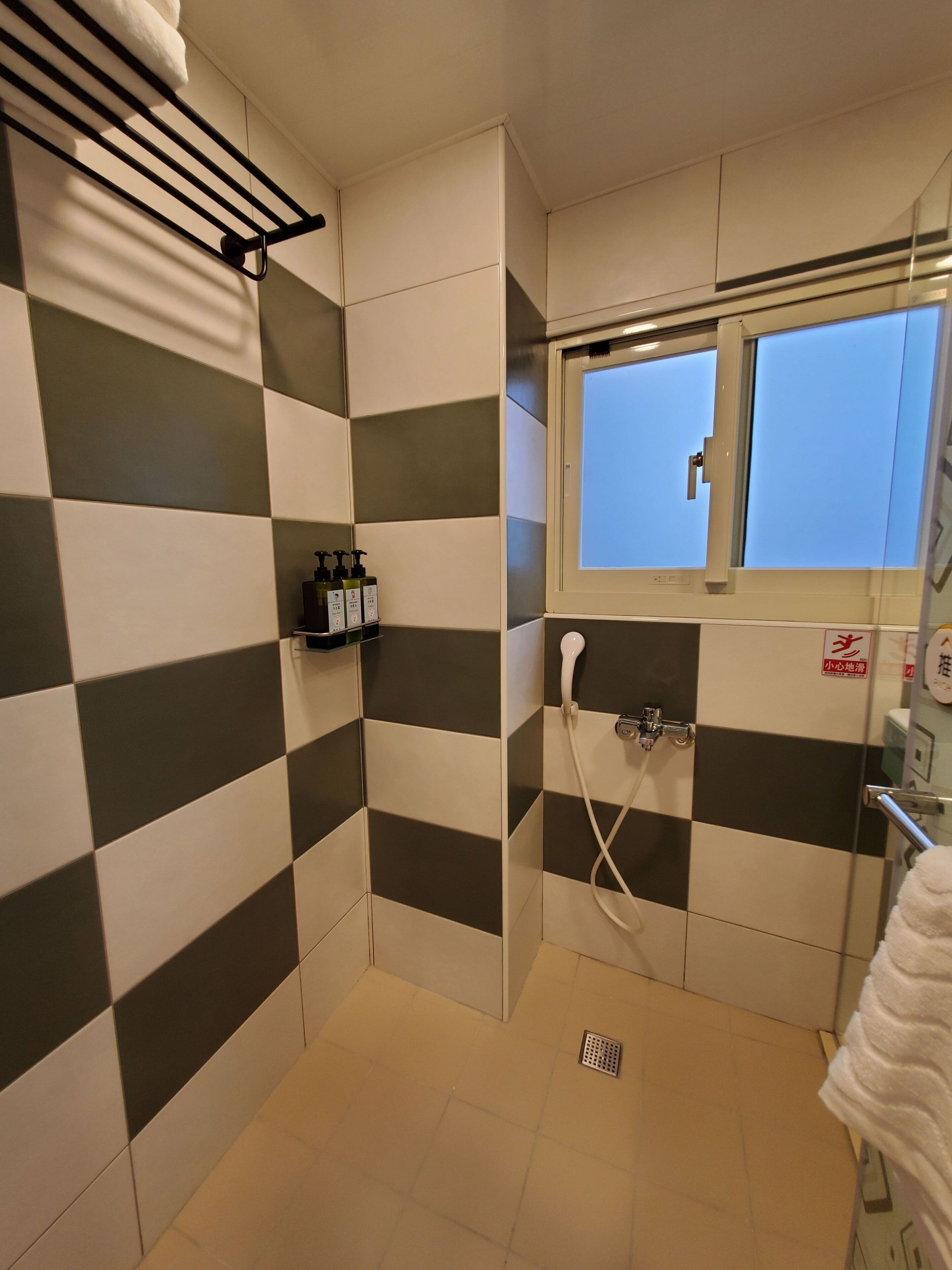 Een kleine betegelde douchekamer met een raam, een handdoekhouder en toiletartikelen aan de muur.