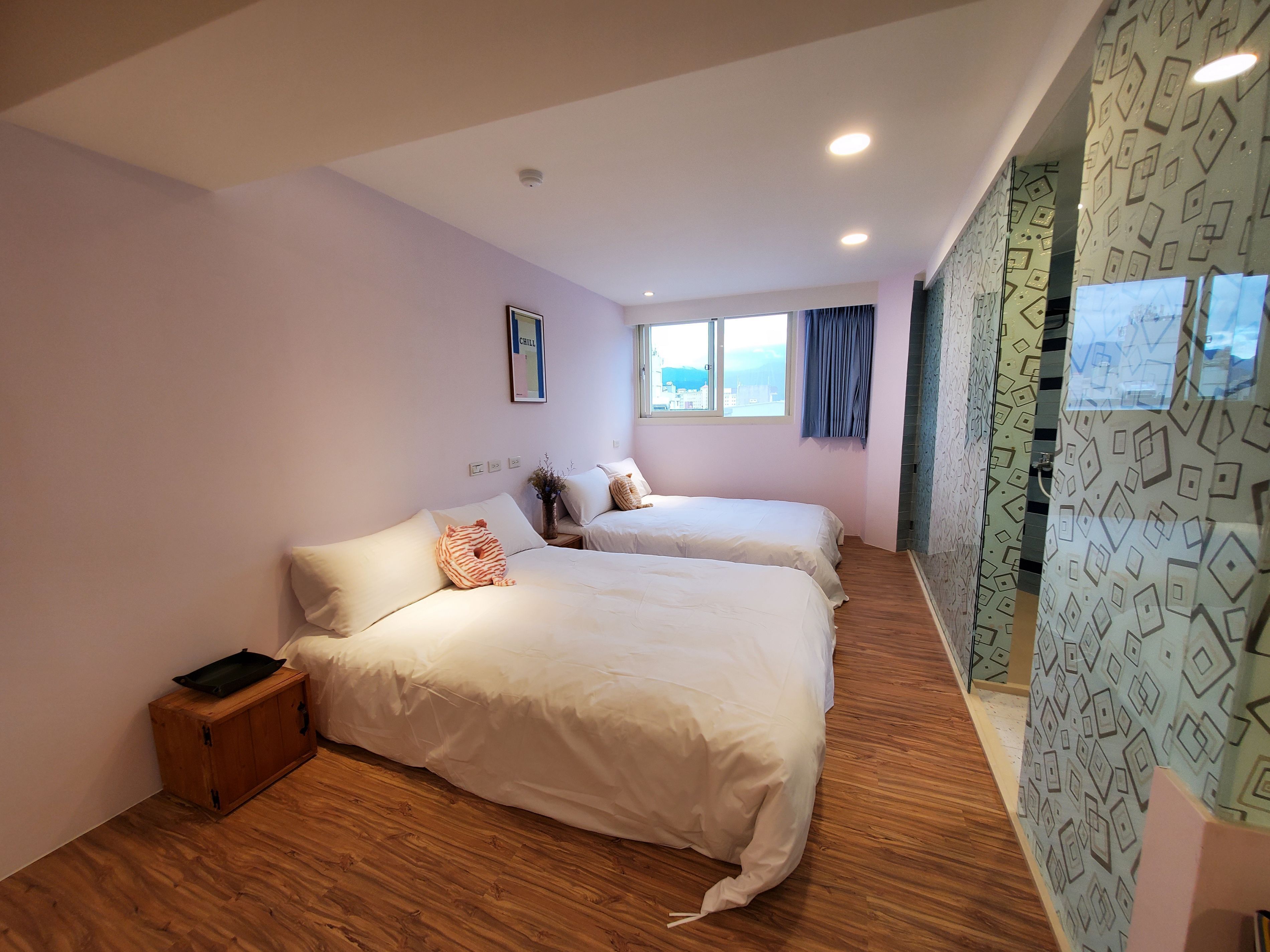 A hotel room with two beds, a small wooden cabinet, a window with a view, and a patterned wall.