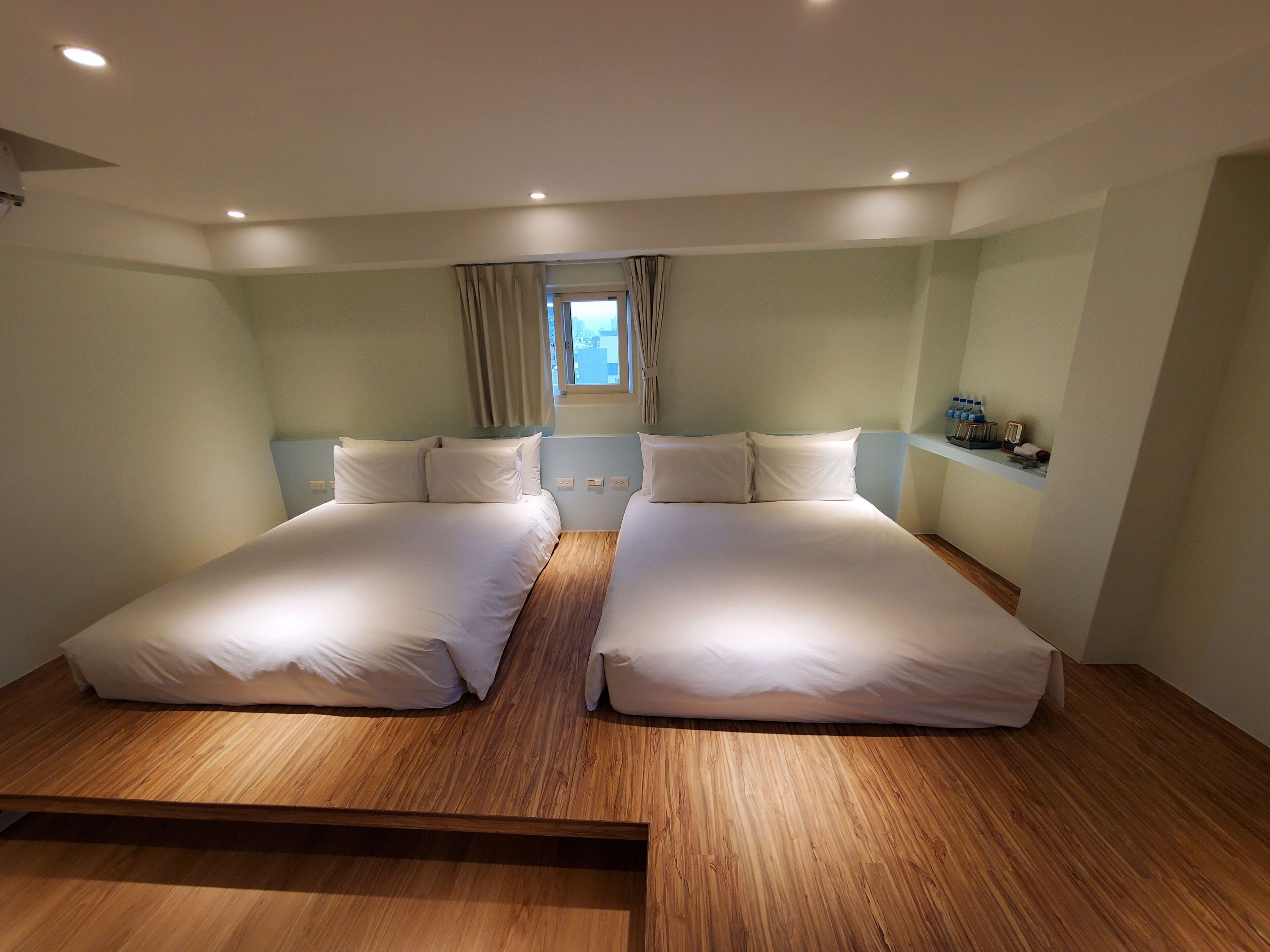 Habitación de hotel con dos camas, piso de madera, una ventana con cortinas y un pequeño estante con objetos.