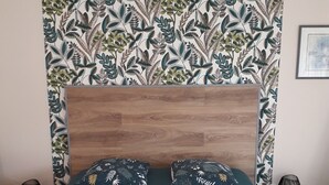 Een net opgemaakt bed met een bedspreide met dessin, twee kussens met bladmotief, een houten hoofdbord en een wandtapijt met een bladpatroon.