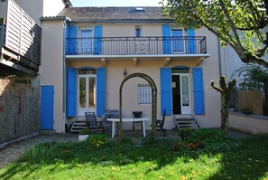 A two-story building with blue shutters and a balcony. There is a white table and chairs in the front yard.