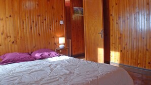 Une pièce aux murs en bois, un lit avec une literie blanche et des oreillers violets, une table de chevet avec une lampe, et une porte donnant sur une autre pièce.