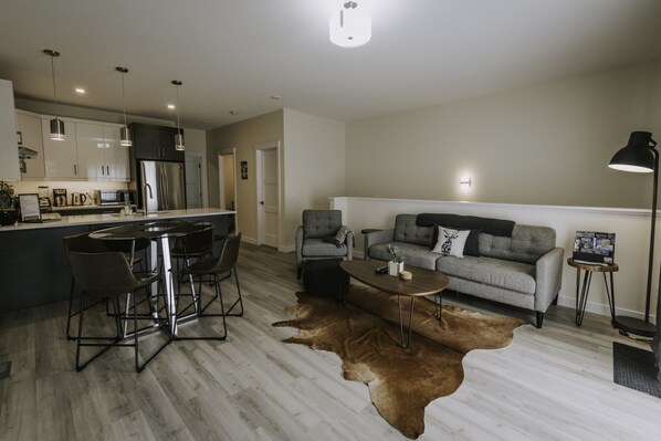 A modern kitchen with stainless steel appliances, a dining area with a round table and chairs, a grey sofa, and a large brown leather rug.