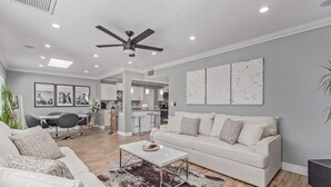 A modern living room with a white sofa, a coffee table, and a ceiling fan. There is a kitchen area in the background with white cabinets and a dining table. - 4 Min 2 Nightlife l HUGE Cabana l Pool & Spa l Golf l BBQ
 (Scottsdale)