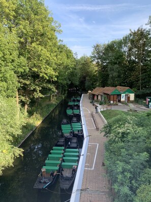Un canal con botes remos amarrados a la orilla, un paseo peatonal y un pequeño edificio con techo verde.