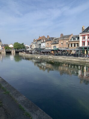 Vista a la orilla del río con edificios, mesas al aire libre y un puente.