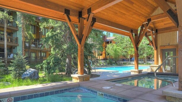 Ein überdachter Poolbereich im Freien mit Holzbalken und Steinstufen, umgeben von üppigem Grün und Blick auf ein mehrgeschossiges Gebäude.