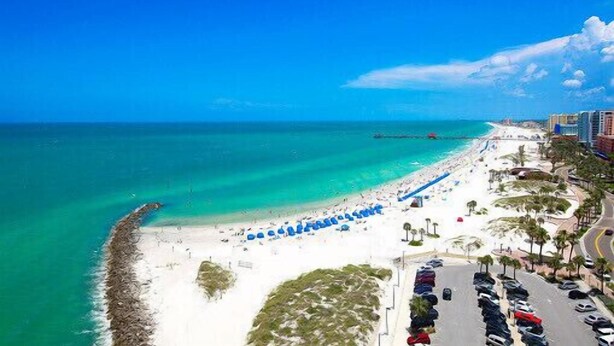 An aerial view of a beach with a white sandy shore, a clear blue sea, and a road lined with parked cars and palm trees. - Beautiful Spacious 3 bedroom home near Clearwater Beach, Pet friendly! (Clearwater)
