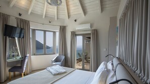 A hotel room with a large bed, a desk with a chair, a TV, and a balcony with a view of the sea. - Villa Mare with Private Pool and Sea Views (Vasiliki)
