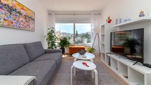 Una sala de estar moderna con un sofá gris, un televisor sobre un mueble blanco, una mesita pequeña y un ventanal con vistas a la ciudad.