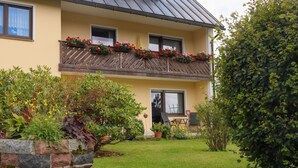 Ein zweistöckiges Gebäude mit einem Balkon, der mit roten Blumen geschmückt ist, einem Steingartenbeet mit verschiedenen Pflanzen und einem gepflegten Rasen.