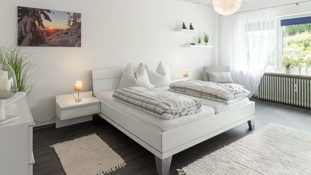 Ein modernes Schlafzimmer mit einem weißen Bett, Nachttischen, einer Hängelampe und Blick auf Grün durch das Fenster.