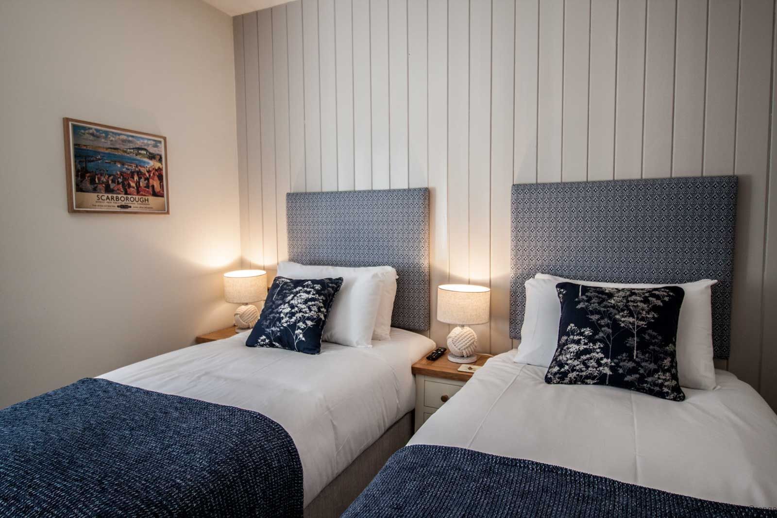A hotel room with two beds, each with a headboard, bedside lamps, and decorative pillows. A framed picture hangs on the wall.