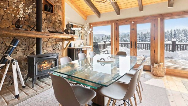 A cozy room with a glass table, chairs, a stone wall, a fireplace, and a view of snow-covered mountains. - 3 BDR Retreat w/ Stunning Mountain & Forest Views! (Breckenridge)