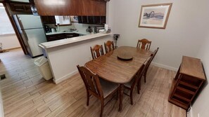A dining area with a wooden table and chairs, a kitchen in the background, and a framed picture on the wall.