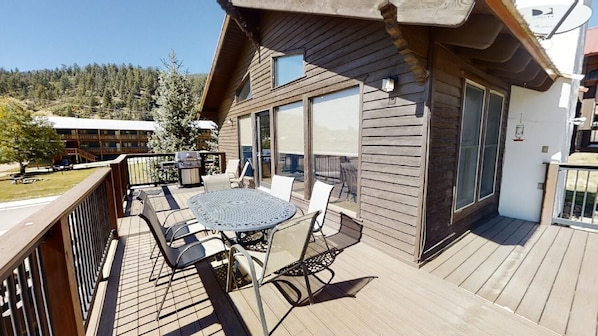A wooden deck with a round table and chairs, large windows, and a view of a forested area.