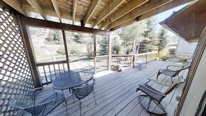 Une terrasse en bois avec une table et des chaises, une balustrade en bois, et une vue sur des arbres et une maison en arrière-plan.