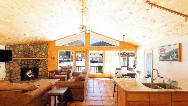 A cozy cabin interior with a stone fireplace, wooden furniture, a kitchen area, and a large window offering a view of the outside. - Ski View Chalet - In Town - New Property - Pet Friendly (Red River)