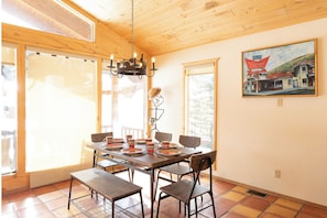 A dining area with a wooden table set for six, surrounded by chairs, a chandelier, and a painting of a building on the wall.