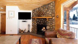 A cozy living room with a stone fireplace, wooden ceiling, and a large window overlooking a snowy landscape. - Ski View Chalet - In Town - New Property - Pet Friendly (Red River)