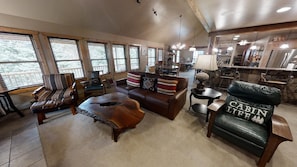 A spacious living room with a large sofa, a coffee table, and a leather chair with a cabin life pillow. There are multiple windows providing natural light and a view of the outdoors.