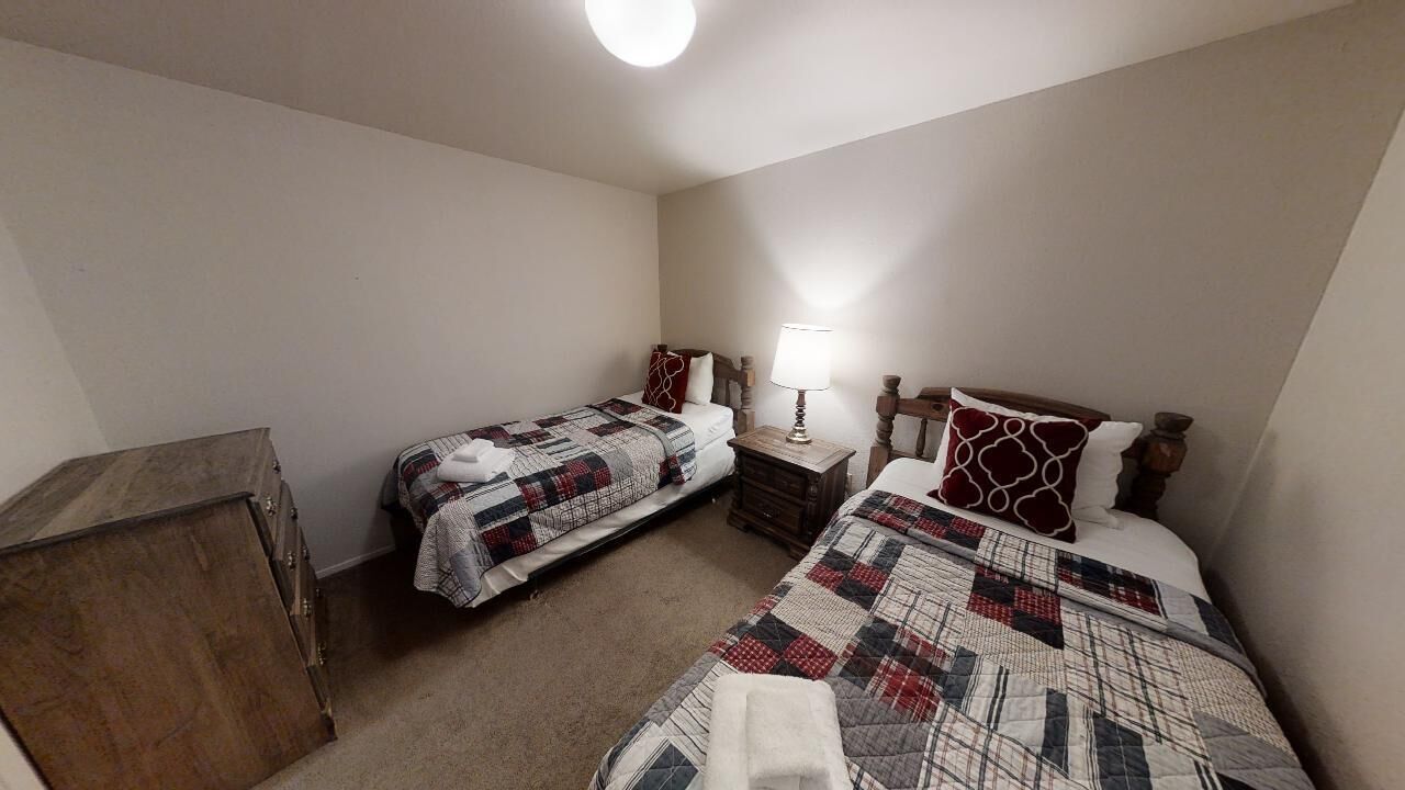 A small, dimly lit room with two single beds, a wooden dresser, and a nightstand with a lamp.