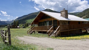 A wooden lodge with a stone chimney, a porch, and a view of mountains.