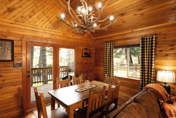 L’intérieur d’un chalet en bois comprend une table à manger, des chaises, un lustre, un canapé et une fenêtre ornée de rideaux à carreaux.