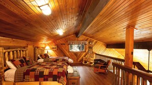 Ein gemütliches Schlafzimmer in einer Holzhütte, mit Holzbalkendecke, zwei Betten mit karierter Bettwäsche, einem Nachttisch, einem Schaukelstuhl und einem Fenster mit Blick.