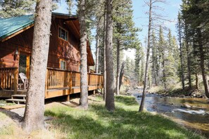 Une cabane en bois dotée d’une terrasse, entourée d’arbres et bordée par un ruisseau.