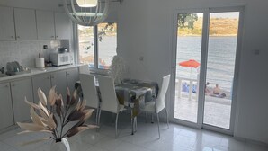 Uma cozinha moderna com armários brancos, uma área de jantar com mesa e cadeiras, e uma grande porta de vidro deslizante que dá para uma varanda com vista para o mar.