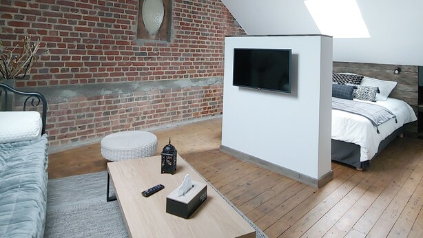 Une chambre à coucher de style « loft » avec des poutres en bois apparentes, un mur en briques, un téléviseur fixé au mur, un lit avec une literie blanche, une table basse en bois et une petite lanterne décorative.