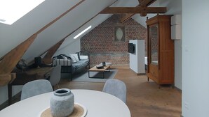 Une pièce de style « loft » avec un plafond en pente, des poutres apparentes, un mur en briques et une lucarne. On y trouve une table ronde avec des chaises, un canapé et un luminaire suspendu.