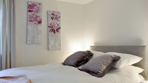 Ein ordentlich bezogenes Bett mit weißen Leinen und zwei Kissen, ein Nachttisch sowie zwei floral verzierte Wandbilder.