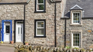 A stone building with two windows and a door, a stone wall, and a small garden area with plants. - A delightful little cottage in a peaceful village location. (New Galloway)