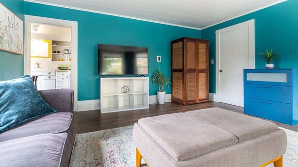 A living room with teal walls, a grey sofa, a beige ottoman, a TV on a white stand, a wooden cabinet, and a blue filing cabinet. - Spunky Multi-Level Unit Near Fremont Seattle (Seattle)