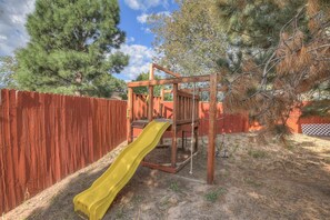 Children can play on the playground structure featuring a slide surrounded by a fence in the backyard.