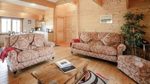 Interior de una cabaña de troncos con dos sofás estampados, una mesa de centro de madera, un comedor y una cocina.