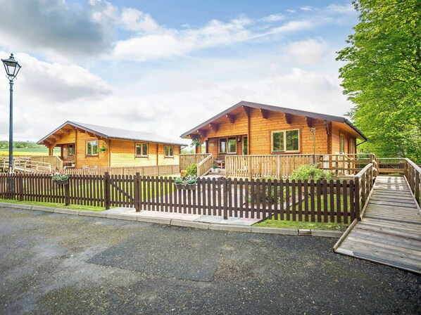 A wooden cabin with a porch and a deck, surrounded by a wooden fence and a paved walkway.