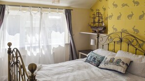 Een slaapkamer met een bed, gordijnen, een kroonluchter, een nachtkastje en een muur met een konijnenprint.