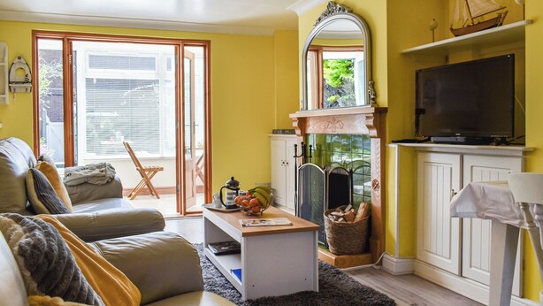 Een woonkamer met een bank, salontafel en een televisie. Er is een open haard met een spiegel erboven, en een raam met jaloezieën.