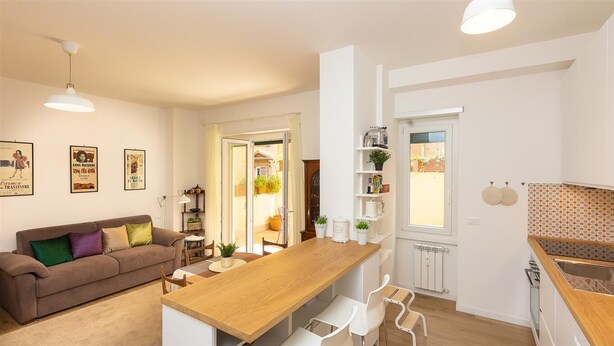 A modern kitchen with a wooden countertop, white chairs, and a dining table. There is a brown sofa with colorful cushions in the living area.
