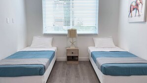 Quarto com duas camas, uma mesinha de cabeceira, um abajur e uma janela com persianas.